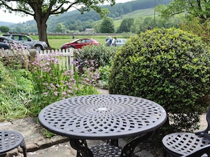 Un jardín con una mesa y sillas metálicas redondas, rodeado de follaje y flores.