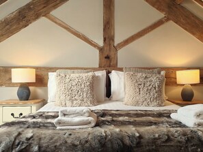 A bedroom with a high ceiling, exposed wooden beams, a bed with fluffy pillows, two bedside lamps, and a fur throw on the bed.