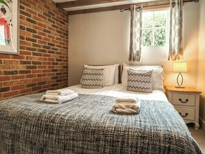 Una habitación con pared de ladrillo, una cama con dos almohadas, una mesita de noche con una lámpara y una ventana con cortinas.