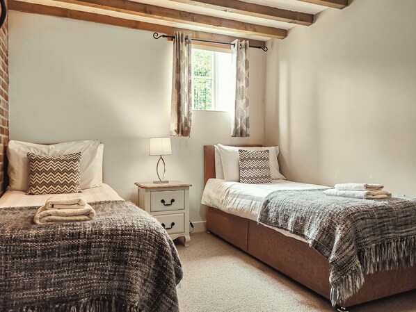 A bedroom with two beds, a nightstand with a lamp, a window with curtains, and a brick wall.