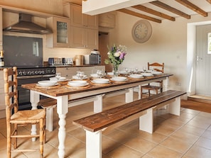 A dining area with a long wooden table set for a meal, a kitchen in the background, and a clock on the wall.