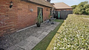 A brick building with a tiled roof, a small patio with a table and chairs, and a garden area with a hedge. - Treacle Cottage at Treacle Den - E5385 (West Hythe, near Romney Marsh)