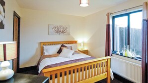 A bedroom with a wooden bed, two bedside lamps, a ceiling light, a window with curtains, and a wall-mounted artwork. - Treacle Cottage at Treacle Den - E5385 (West Hythe, near Romney Marsh)