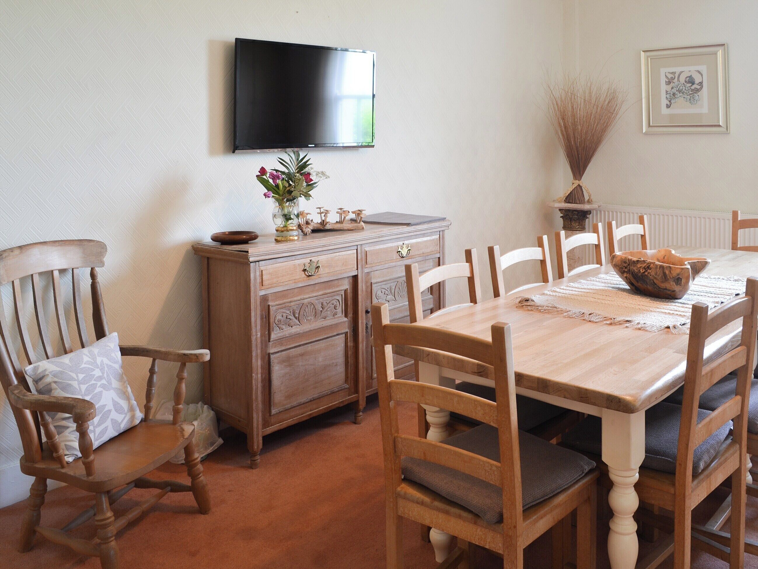Un coin repas comprenant une table en bois, des chaises, un téléviseur fixé au mur et un vase décoratif avec des fleurs.