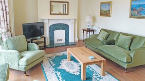 Een woonkamer met een open haard, een groene bank, fauteuil, televisie en een kroonluchter.