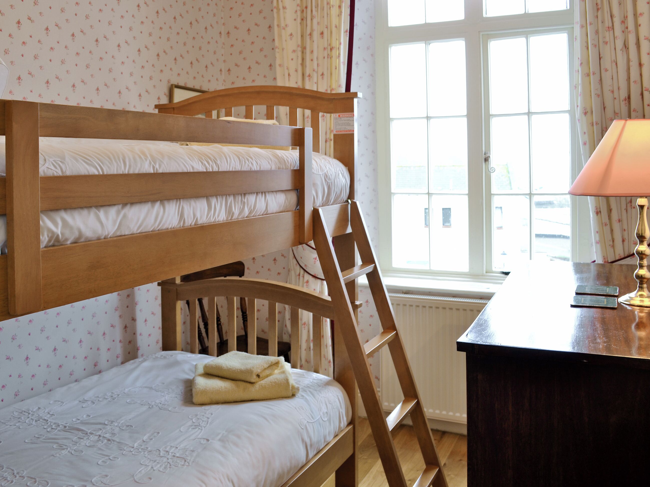 A bunk bed with a wooden ladder, a desk with a lamp, and a window with curtains.