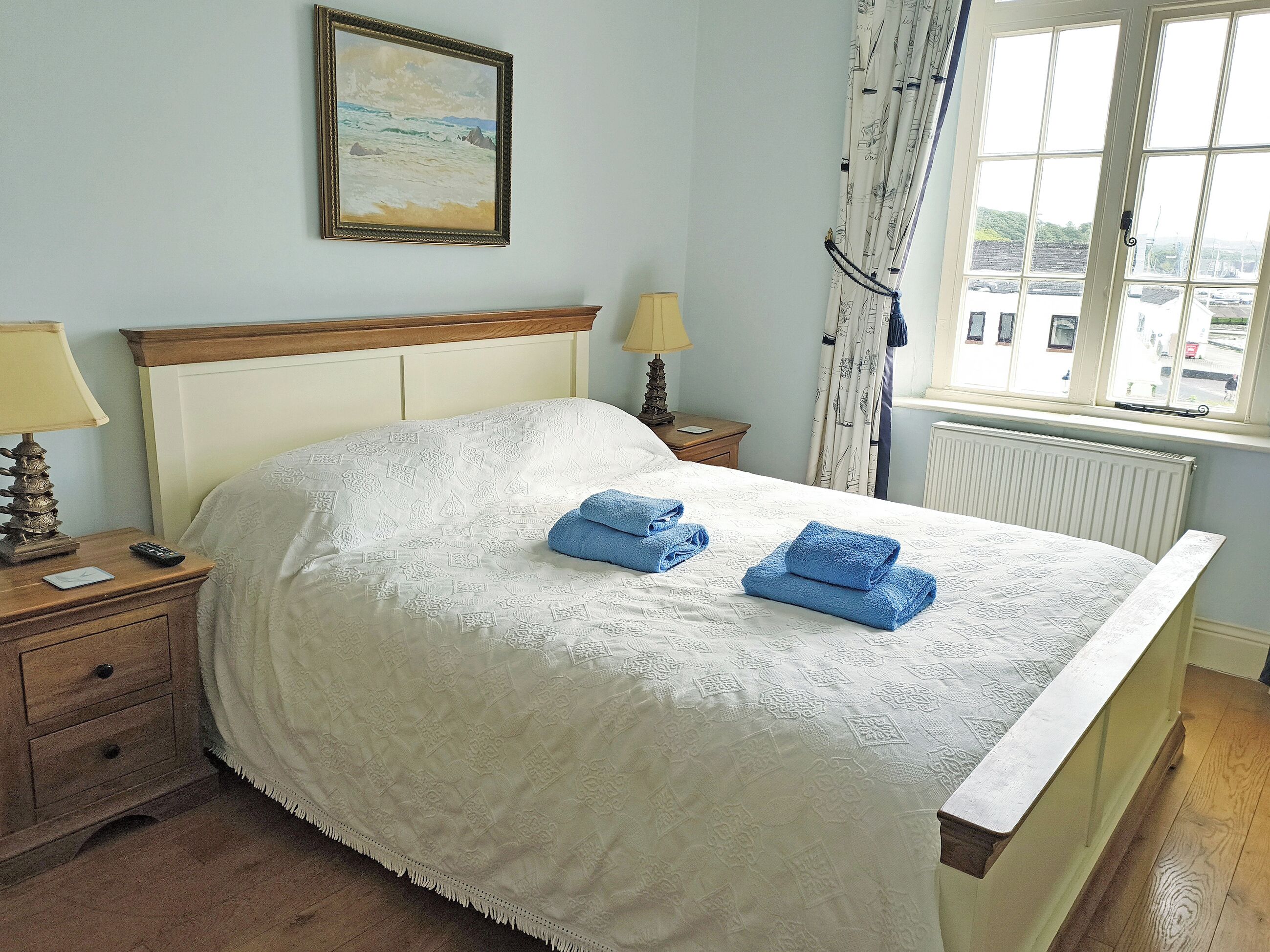 A neatly made bed with a white bedspread and two folded blue towels, a bedside lamp, a nightstand with a remote control, and a framed picture on the wall.