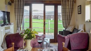 A dining area with a table set for two, a television in the background, and a view of a green landscape through large windows. - 2 bedroom accommodation in Hooe, Battle (Hooe, Battle)