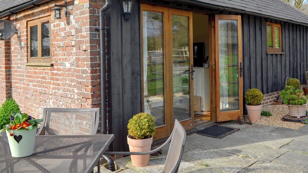A small brick building with a dark wooden extension, large glass doors, and a patio area with outdoor furniture and potted plants. - 2 bedroom accommodation in Hooe, Battle (Hooe, Battle)