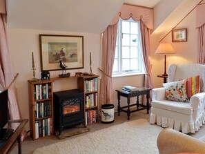 A cozy living room with a fireplace, bookshelves, a white armchair, and a window with curtains. - 1 bedroom accommodation in Fylingthorpe, near Whitby (Fylingthorpe, near Whitby)