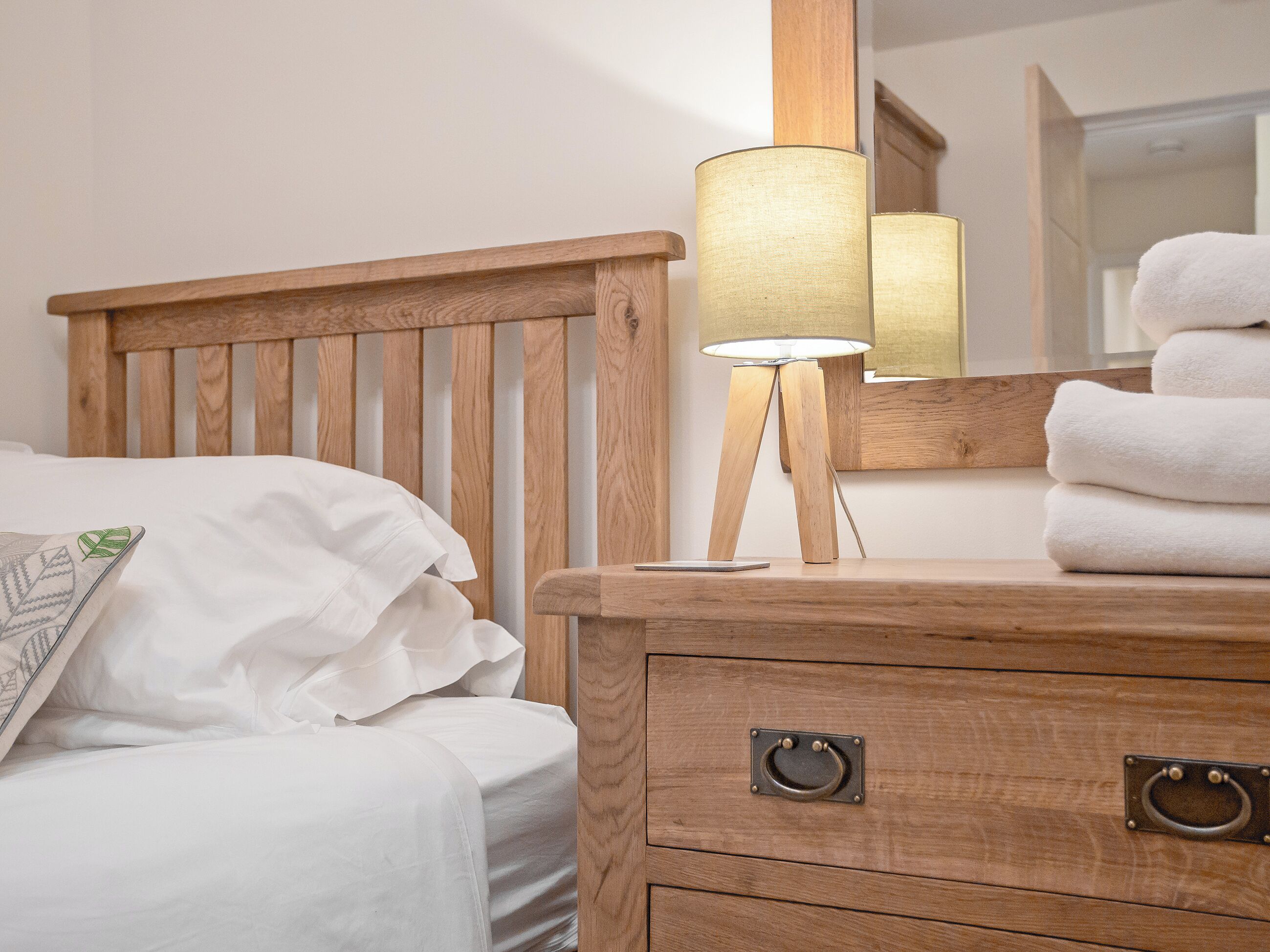 A wooden bed with white bedding, a bedside lamp, a mirror, and a stack of towels.
