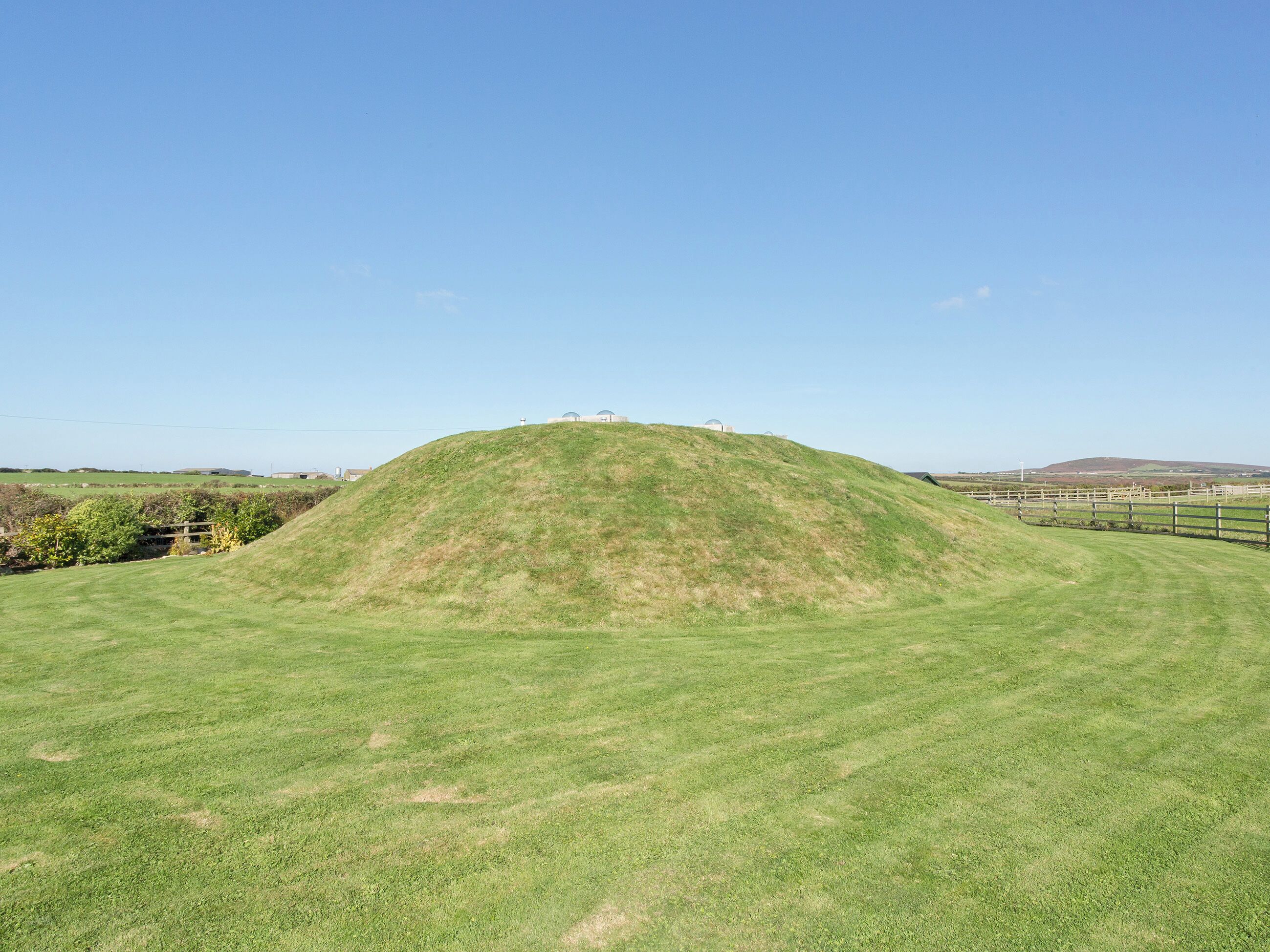 A grassy hill with a small building on top, surrounded by open fields and a clear sky.