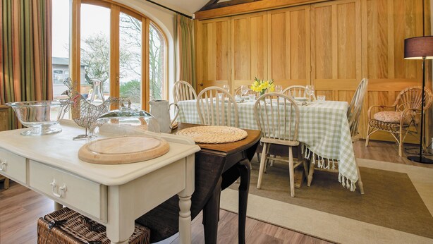 A dining area with a table set for four, wooden paneled walls, and a large window with curtains. - The Stables at Weedon Hill Farm (Weedon, near Daventry)