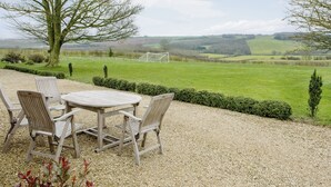 A gravel patio with a round table and four chairs, overlooking a green field and hills. - The Stables at Weedon Hill Farm (Weedon, near Daventry)