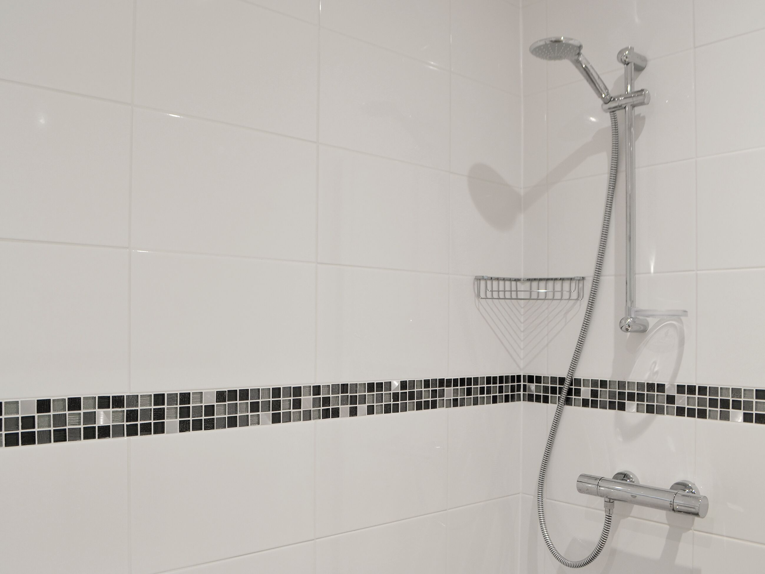 Ein Badezimmer mit weißen Fliesen, einem schwarz-weißen Mosaikstreifen und einer chromfarbenen Duscharmatur.