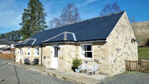 Un edificio de piedra con techo oscuro, un letrero que dice "No 2 Railway Cottages" y asientos al aire libre.