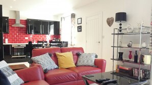 Una sala de estar con un sofá rojo, una mesa de centro de cristal y una cocina al fondo con baldosas rojas.