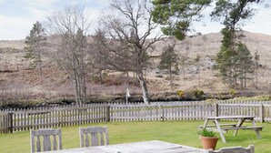 A rustic outdoor seating area with a wooden table and chairs, surrounded by a wooden fence, set against a backdrop of trees and hills. - 2 bedroom accommodation in Rosehall, near Lairg (Rosehall, near Lairg)