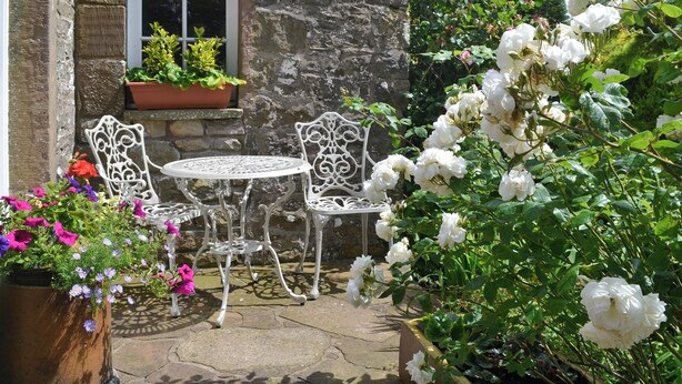Un bâtiment en pierre avec une fenêtre, deux chaises en métal blanches, une petite table ronde et des plantes en pot avec des fleurs.