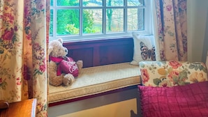 Un siège de fenêtre agrémenté d’un ours en peluche, de rideaux à motifs floraux et d’un coussin orné de motifs de papillons.