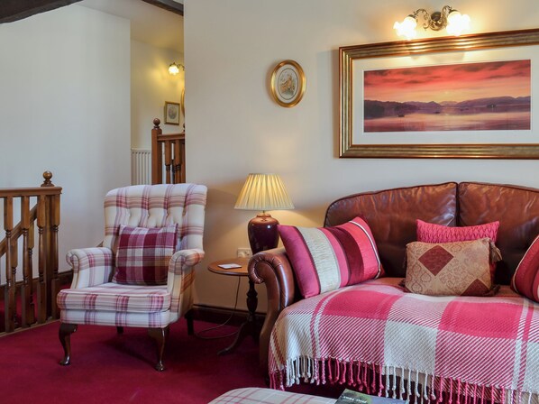 A living room with a plaid armchair, a leather sofa, a small round table with a lamp, and a framed painting on the wall.