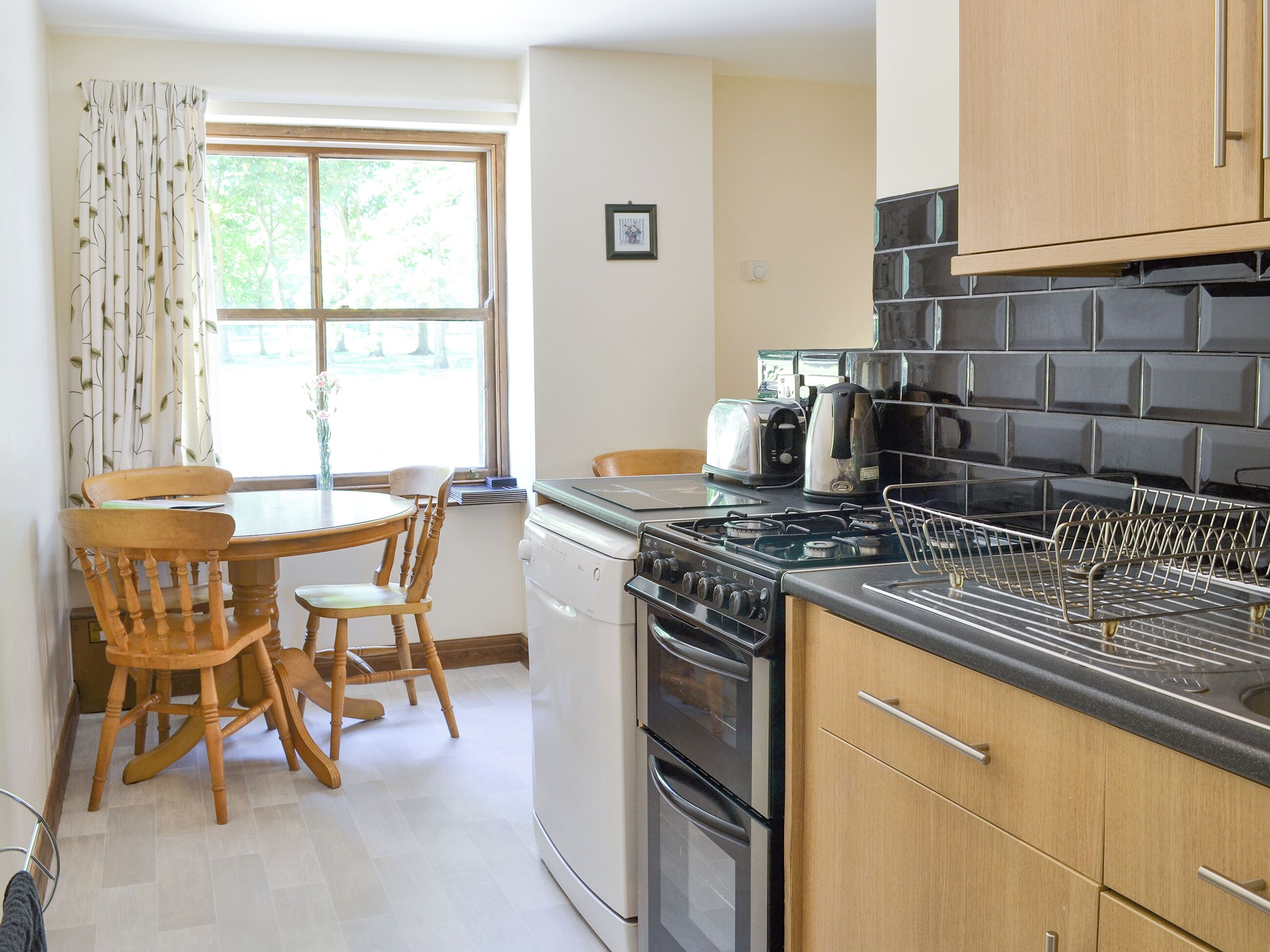 A kitchen with a stove, oven, and a drying rack. There is a window with curtains, a table with chairs, and a framed picture on the wall.