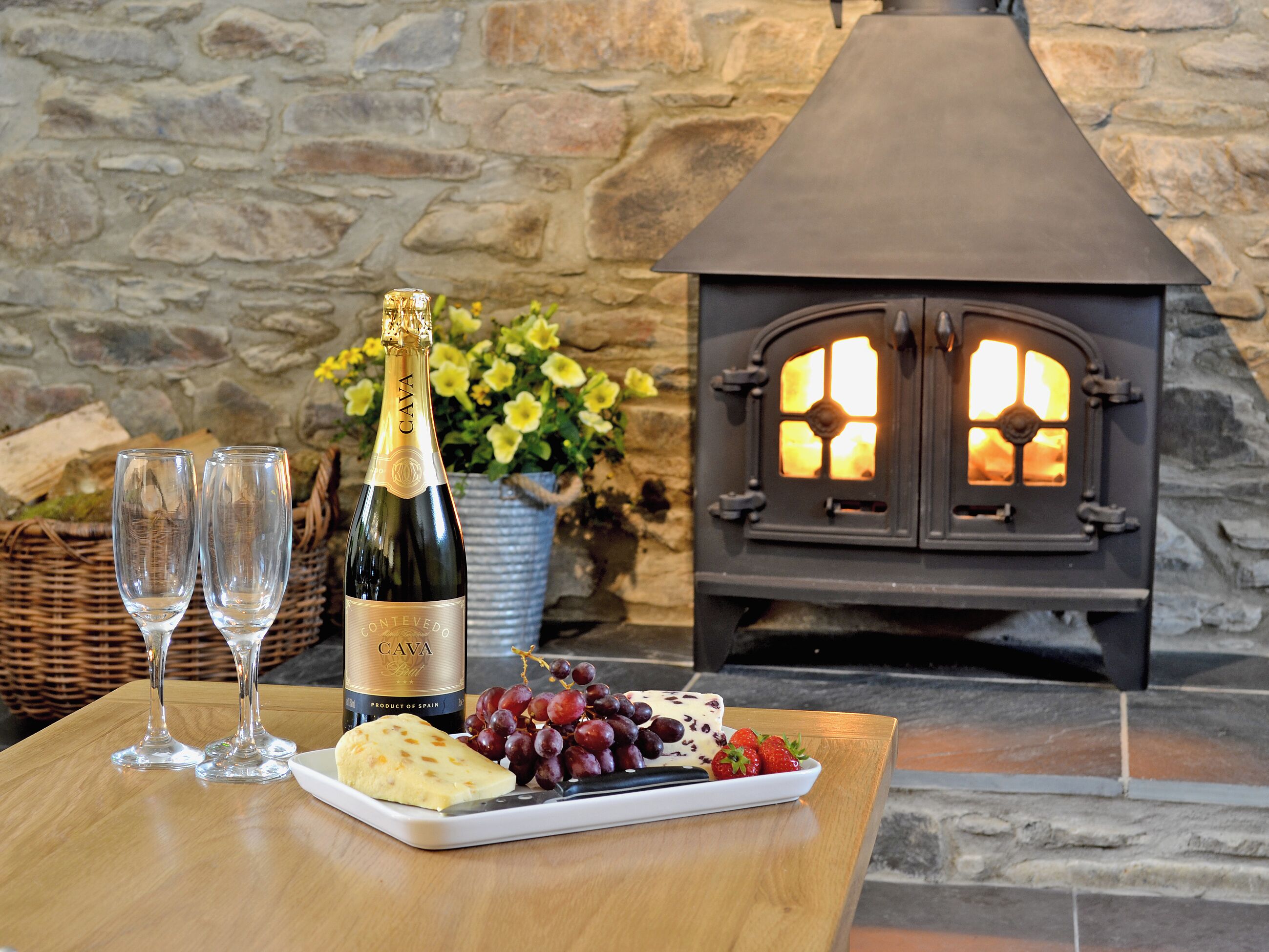 A table with a bottle of Cava, two glasses, a plate of cheese, grapes, and strawberries, and a wood-burning stove in the background.