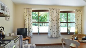 A cozy kitchen with a stove, kettle, and a television. There are striped chairs and a dining table. Large windows with curtains offer a view of greenery outside.