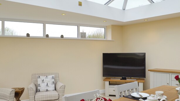 Una habitación amplia con techo alto, ventanas grandes, televisor de pantalla plana, una mesa de comedor con tetera y tazas, y un rincón de estar acogedor con un sillón estampado.