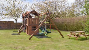 Una estructura de juego de madera con un tobogán y columpios, una mesa de picnic y un muro de ladrillo al fondo.