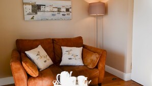 A brown leather sofa with two cushions, a white coffee table with a teapot and cups, a floor lamp, and a painting of a coastal scene on the wall.