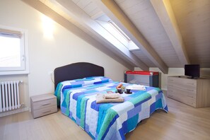 Une chambre avec un plafond en pente, un lit recouvert d’une couette bleue et blanche, une table de chevet avec une lampe, une commode et un téléviseur posé sur un meuble.