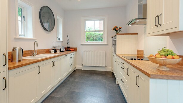 A modern kitchen with white cabinets, a wooden countertop, a large clock, and a window providing natural light. - 3 bedroom accommodation in Filey (Filey)