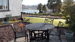 Pátio externo com mesa redonda de metal e quatro cadeiras, com vista para um lago e montanhas.