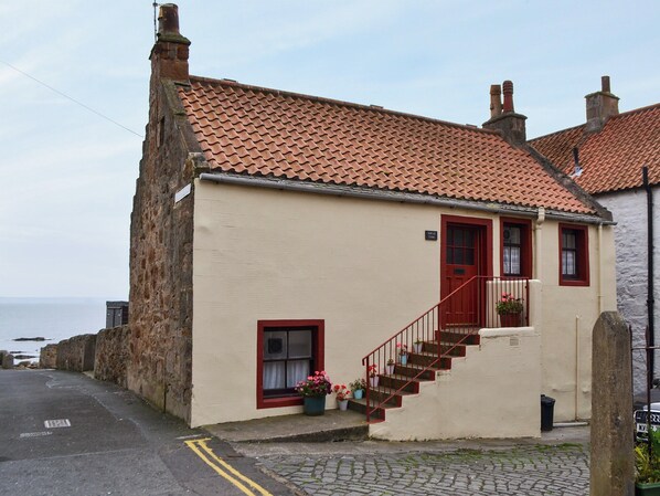 A two-story building with a red-tiled roof, a red door, and a staircase leading to the second floor. There are potted plants on the ground floor.
