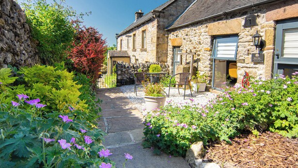Un bâtiment en pierre avec un chemin également en pierre menant à une porte, le tout entouré de verdure et de fleurs.