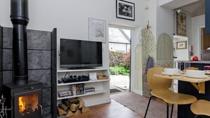 Un salon moderne avec un poêle à bois noir, un téléviseur à écran plat posé sur une étagère blanche, un coin repas avec une table et des chaises en bois, et une vue sur le jardin à travers une porte vitrée.