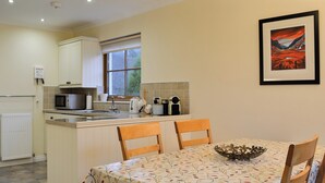 Une cuisine avec un coin repas, comprenant une table recouverte d’une nappe à motifs de feuilles, des chaises en bois, une bouilloire, un grille-pain et un tableau encadré accroché au mur.