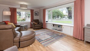 A modern living room with a leather sofa, a brown armchair, a patterned rug, and a bookshelf by a large window with pink curtains.