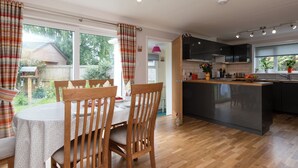 A modern kitchen with a central island, wooden dining table with chairs, and a view of the garden through large windows.
