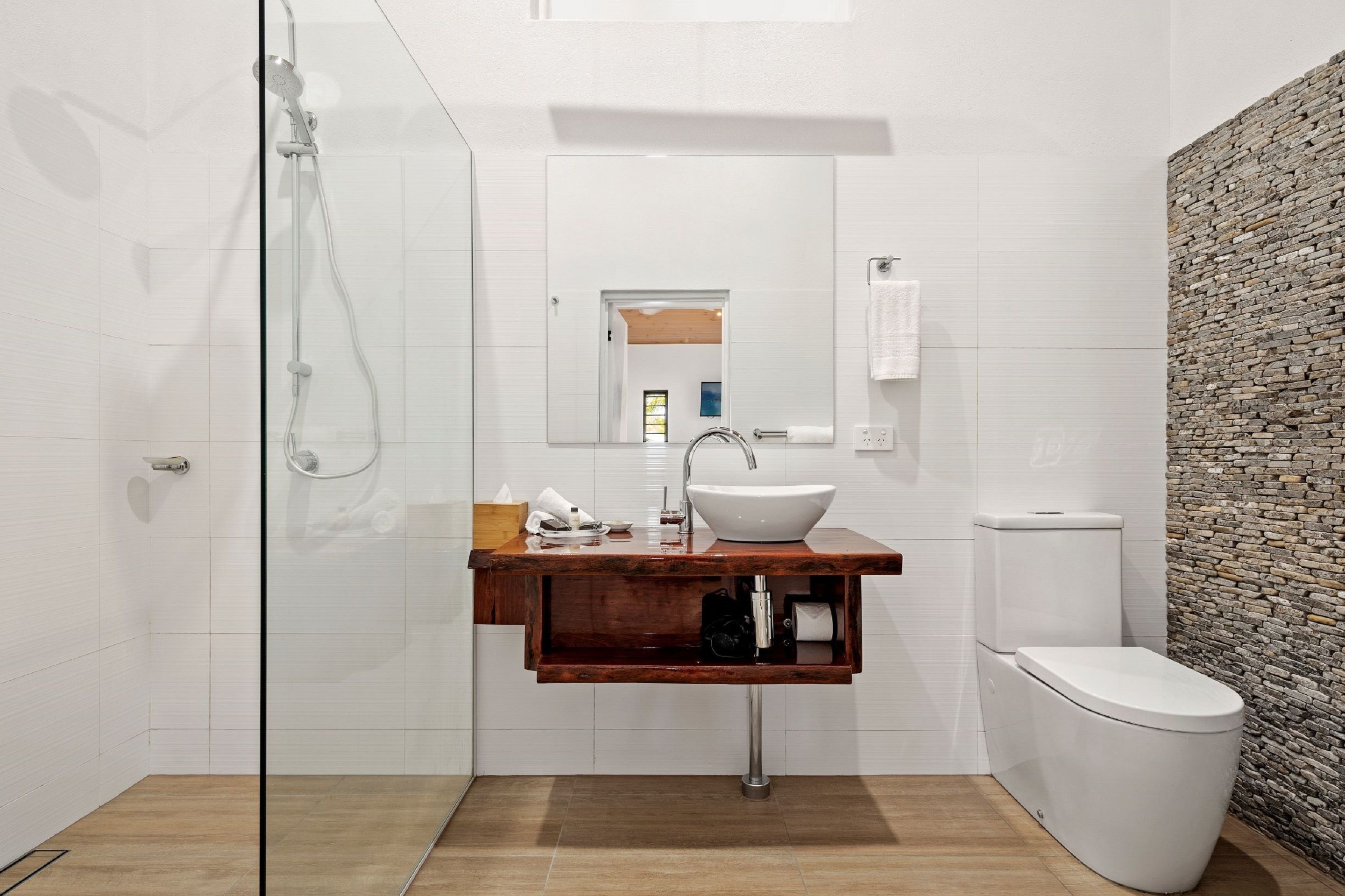 Un baño moderno con cabina de ducha de vidrio, tocador de madera con lavabo blanco y pared con textura de piedra.