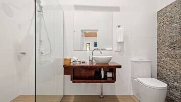 Un baño moderno con cabina de ducha de vidrio, tocador de madera con lavabo blanco y pared con textura de piedra.