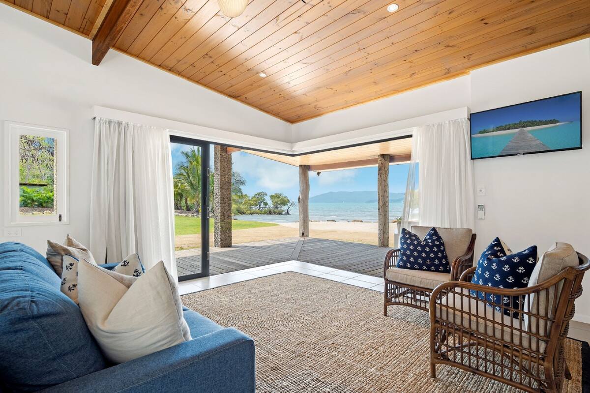 Uma sala de estar espaçosa com teto de madeira, uma janela ampla com vista para a praia, um sofá azul com almofadas náuticas e uma poltrona de vime com almofadas combinando.