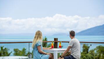 Two people seated at a table with drinks, overlooking a scenic view of the ocean and mountains.