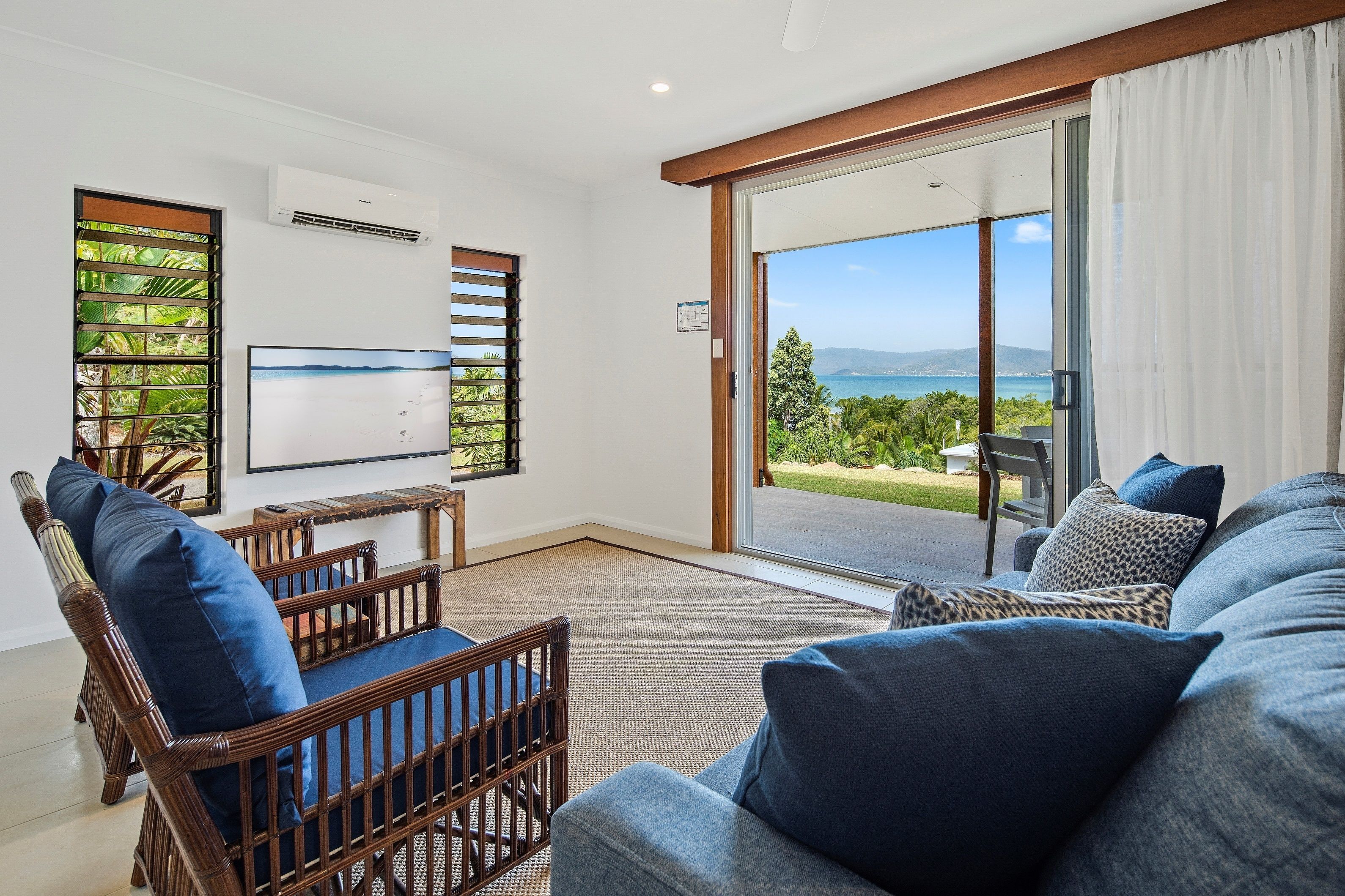 A modern living room with a large sofa, a wooden chair, and a flat-screen TV. There is a sliding glass door leading to a balcony with a view of the ocean.