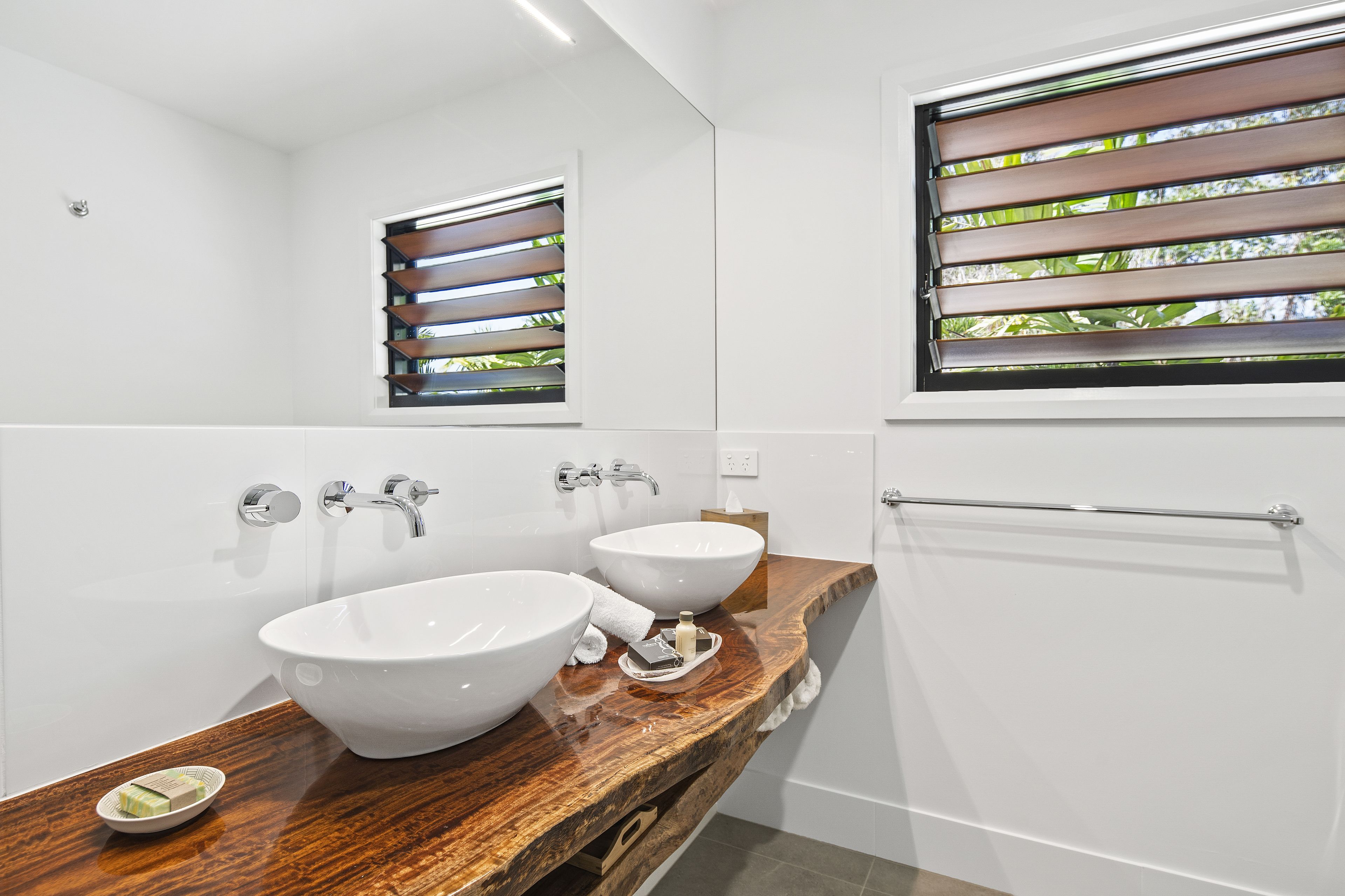 Un bagno moderno con due ampi lavandini bianchi, un mobile contenitore in legno, un porta asciugamani e una finestra con persiane.
