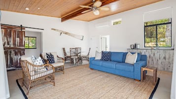 Un vaste espace de vie agrémenté d’un canapé bleu, de fauteuils en osier, d’un plafond en bois et d’une grande fenêtre.