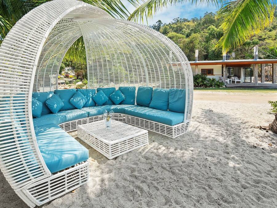 A white wicker lounge chair with blue cushions on a sandy beach, a white coffee table, and a building in the background.