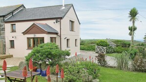 A two-story house with a sloped roof, a garden with red flowering plants, and a view of the countryside. - 2 bedroom accommodation in Pendeen (Pendeen)
