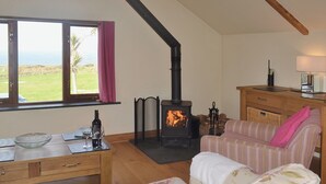 A cozy living room with a fireplace, a sofa, a dining table with a bowl and glasses, and a view of the outdoors. - 2 bedroom accommodation in Pendeen (Pendeen)